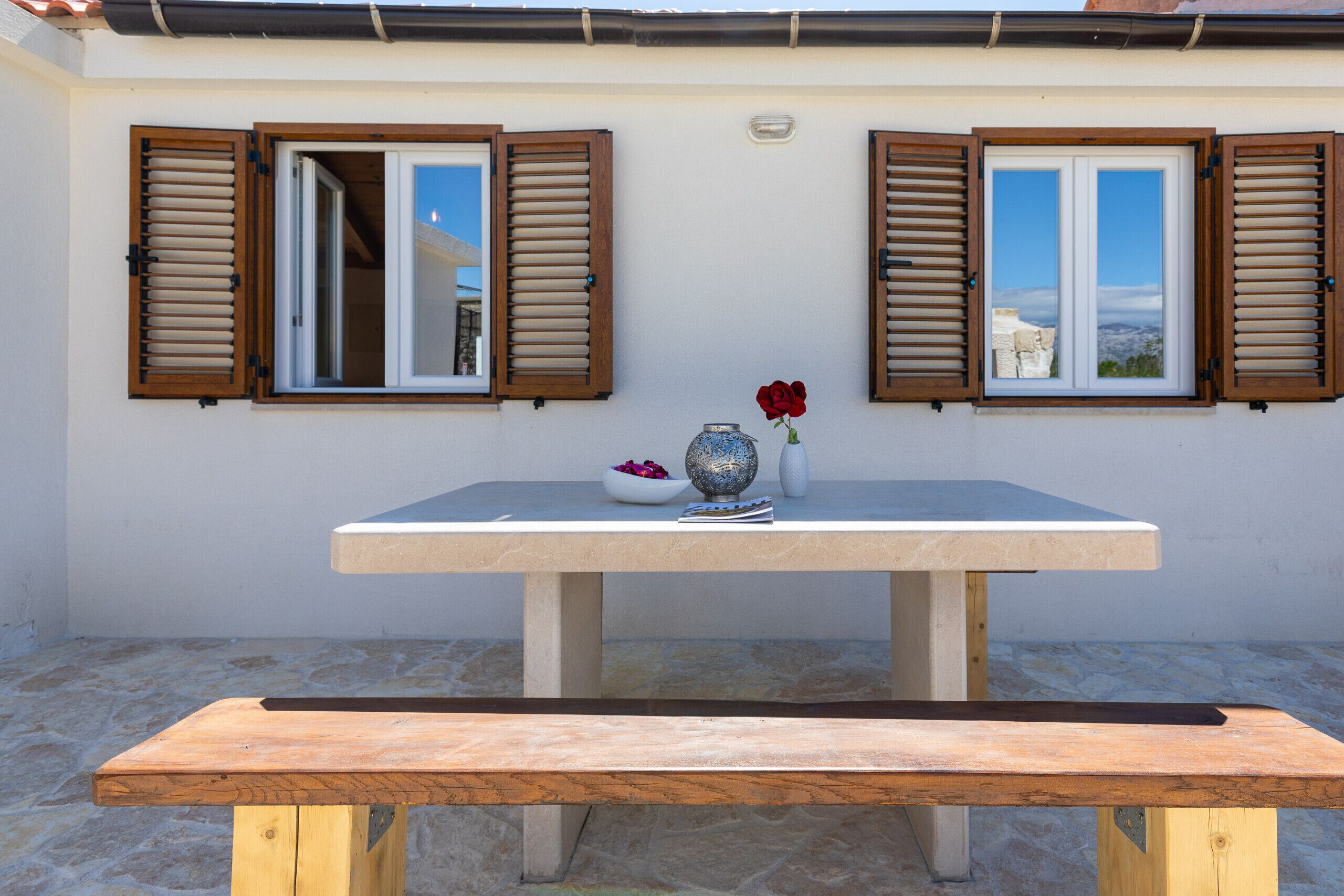 A table with a vase and a book, two windows with shutters, and a bench.