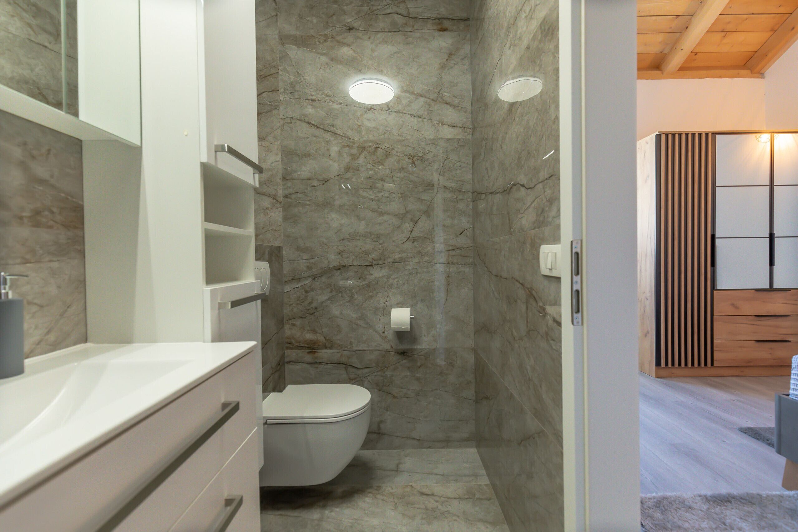 A bathroom with a marble wall, a toilet, a white vanity, and a wooden ceiling.
