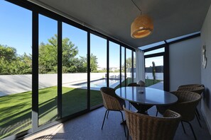 Ein moderner Essbereich mit einem großen Tisch, Korbstühlen und Blick auf einen Swimmingpool und Grünflächen.