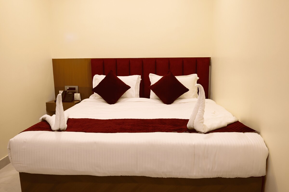 Habitación de hotel con una cama bien hecha, cabecera de madera, mesita de noche con un teléfono y una botella, y una manta roja.