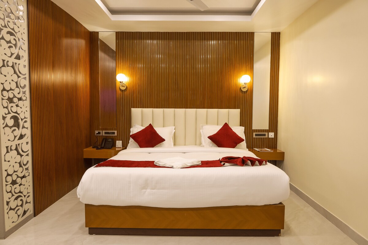 Una cama grande con sábanas blancas, dos almohadas rojas y una manta roja. Hay dos mesitas de noche con lámparas y un teléfono. La habitación tiene paneles de madera en las paredes y una lámpara en el techo.