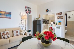 Ein Wohnzimmer mit einer Couch, einem runden Tisch mit Blumenvase, einer Küchenzeile mit Kühlschrank und Schränken sowie einem durchgehenden Türrahmen einsehbaren Badezimmer.