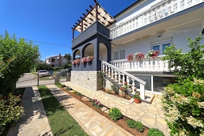 Ein zweistöckiges Gebäude mit einem Steinweg, einer Holzpergola und einem weißen Balkon mit Blumenkästen.