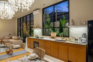 A modern kitchen with a large window, wooden cabinets, and a dining area with a sofa and table set. - Feeels Villas 2 (Badung)