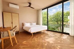 A bedroom with a bed, a ceiling fan, an air conditioner, a wooden table, wicker chairs, and a large window with a view of greenery. - Feeels Villas 2 (Badung)