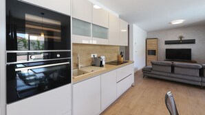 Una cocina moderna con horno integrado, un lavamanos y una encimera. Hay un sofá gris y un televisor en la sala de estar.