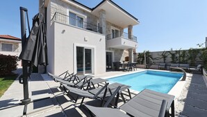 Una casa de dos pisos con piscina, mobiliario exterior y un patio cubierto.