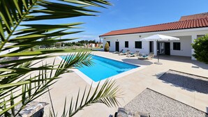 En swimmingpool med solstolar och ett parasoll, en vit byggnad med ett rött tak och palmer.