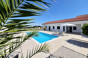 Ein Swimmingpool mit Liegestühlen und Sonnenschirm, ein weißes Gebäude mit rotem Dach und Palmen.
