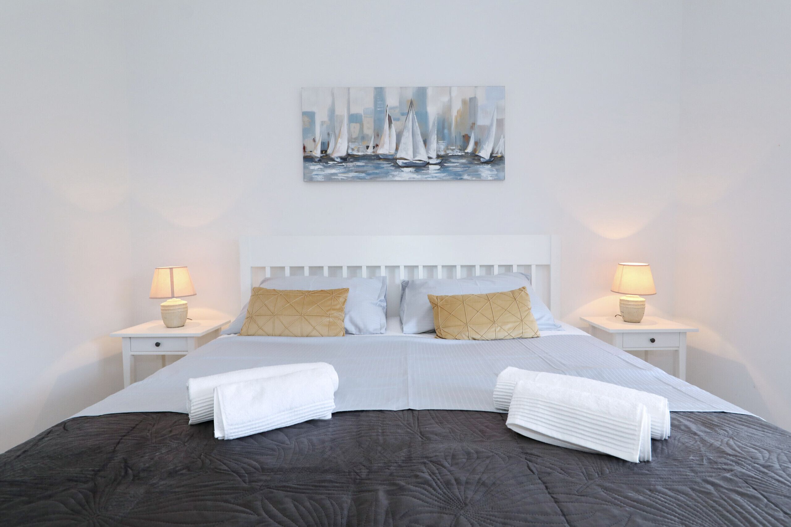 Un lit bien fait, recouvert d’une couverture sombre, deux tables de chevet avec des lampes et un tableau représentant des voiliers accroché au mur.