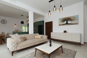 Ein Wohnzimmer mit einer Couch, einem Couchtisch, Pendelleuchten, einem Gemälde und einem Essbereich im Hintergrund.
