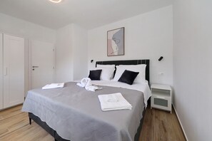 Ein ordentlich bezogenes Bett mit einer grauen Tagesdecke, zwei schwarzen Kissen und einem auf der Seite gefalteten weißen Handtuch.