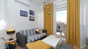 Ett modernt vardagsrum med en soffa, ett soffbord och ett sidobord med en lampa. Det finns gardiner, ett fönster och en väggmonterad luftkonditioneringsanläggning.
