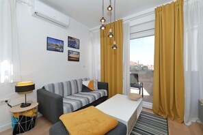 A modern living room with a sofa, a coffee table, and a side table with a lamp. There are curtains, a window, and a wall-mounted air conditioner. - Apartment Bajana in Central Location (Zadar)