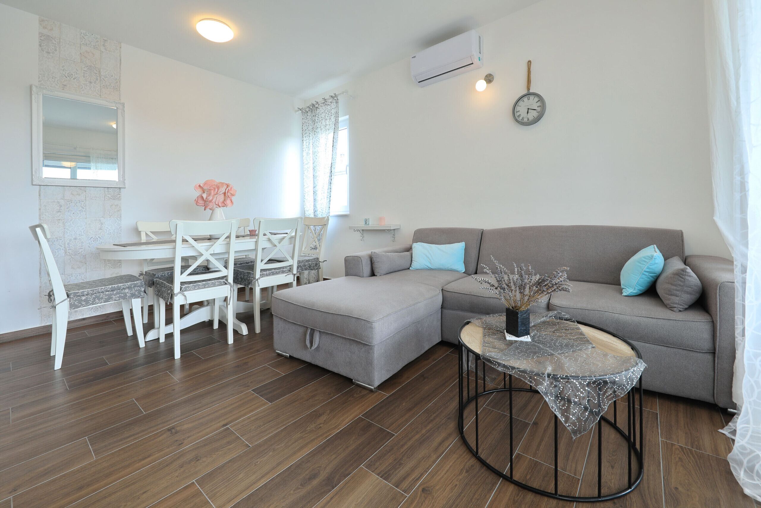 Uma sala de estar com sofá seccional cinza, uma mesa redonda pequena, uma mesa de jantar branca com cadeiras, um relógio na parede e um aparelho de ar condicionado.