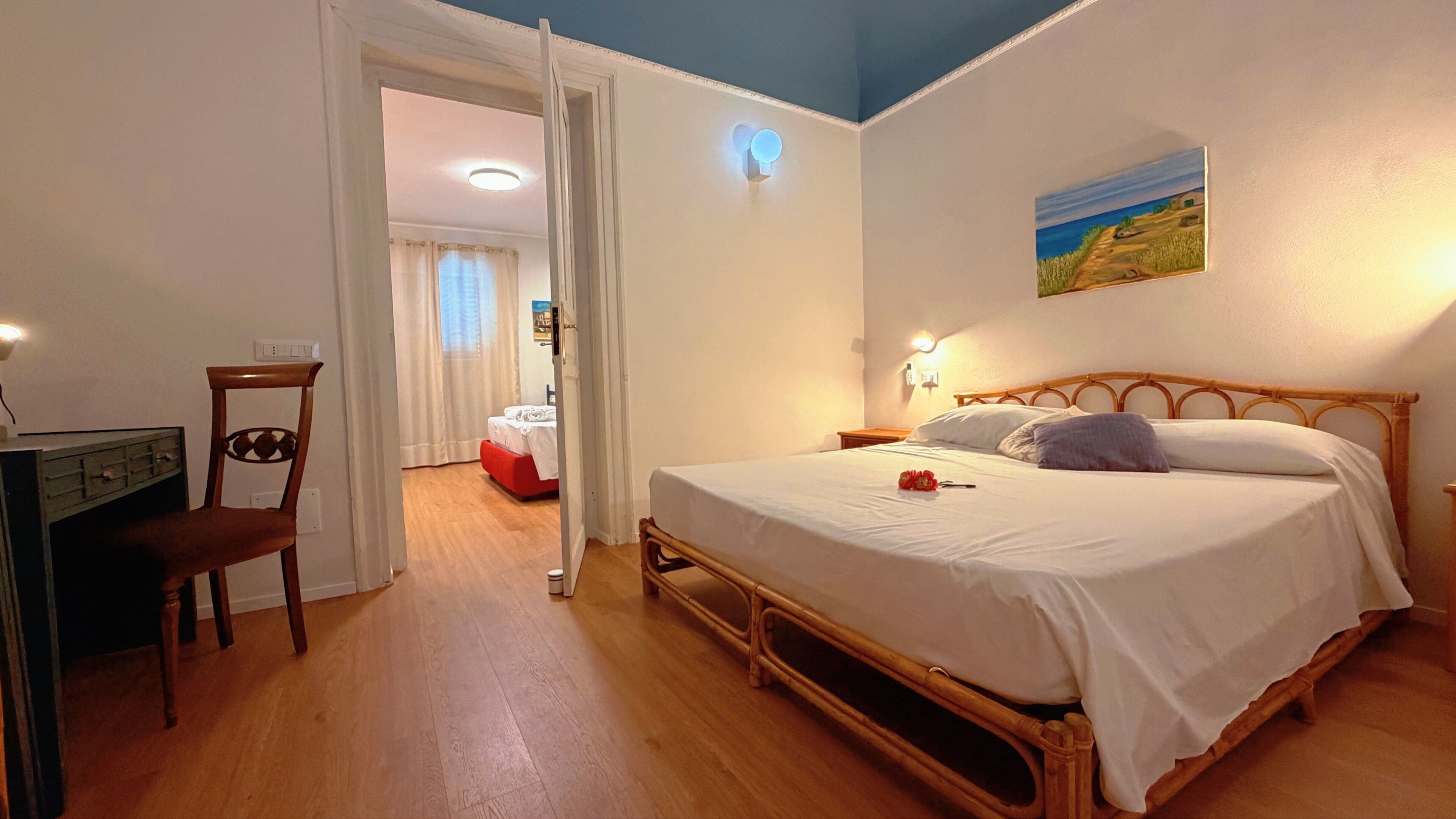 Une chambre avec un lit en bois, un bureau et une chaise. Il y a un tableau accroché au mur et une porte qui donne sur une autre pièce.