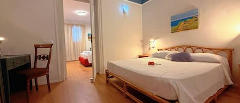 A bedroom with a wooden bed, a desk, and a chair. There is a painting on the wall and a door leading to another room.
