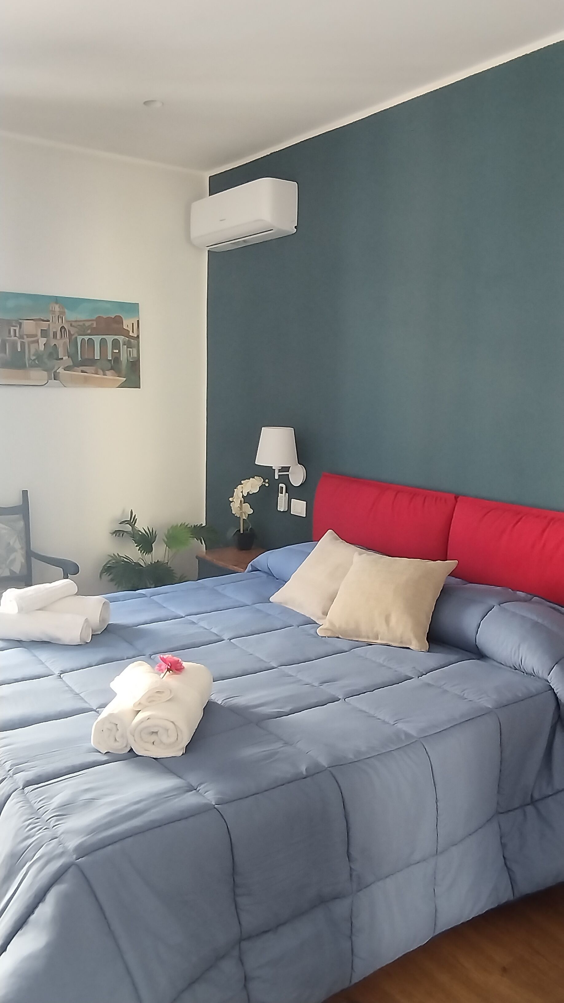A bedroom with a blue bedspread, a red headboard, a wall-mounted air conditioner, a bedside lamp, and a painting of a cityscape.
