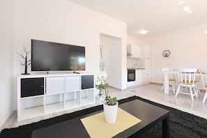 Ein modernes Wohnzimmer mit Fernseher, weißen Schränken, einem Essbereich mit weißen Stühlen und einer Topfpflanze.