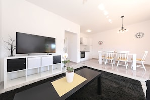 Ein modernes Wohnzimmer mit Flachbildfernseher, Couchtisch mit Pflanze, Essbereich mit weißen Stühlen und einer Küche im Hintergrund.