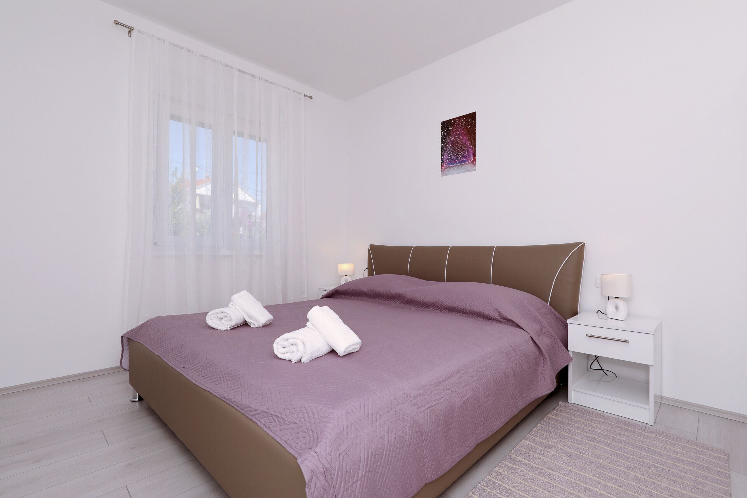 Un dormitorio con una cama, una mesita de noche, una lámpara y una ventana con cortinas translúcidas.