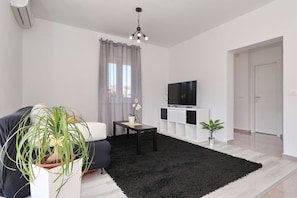 Ein modernes Wohnzimmer mit einer Couch, einem schwarzen Teppich, einem Fernseher auf einem weißen Schrank und einer Topfpflanze.