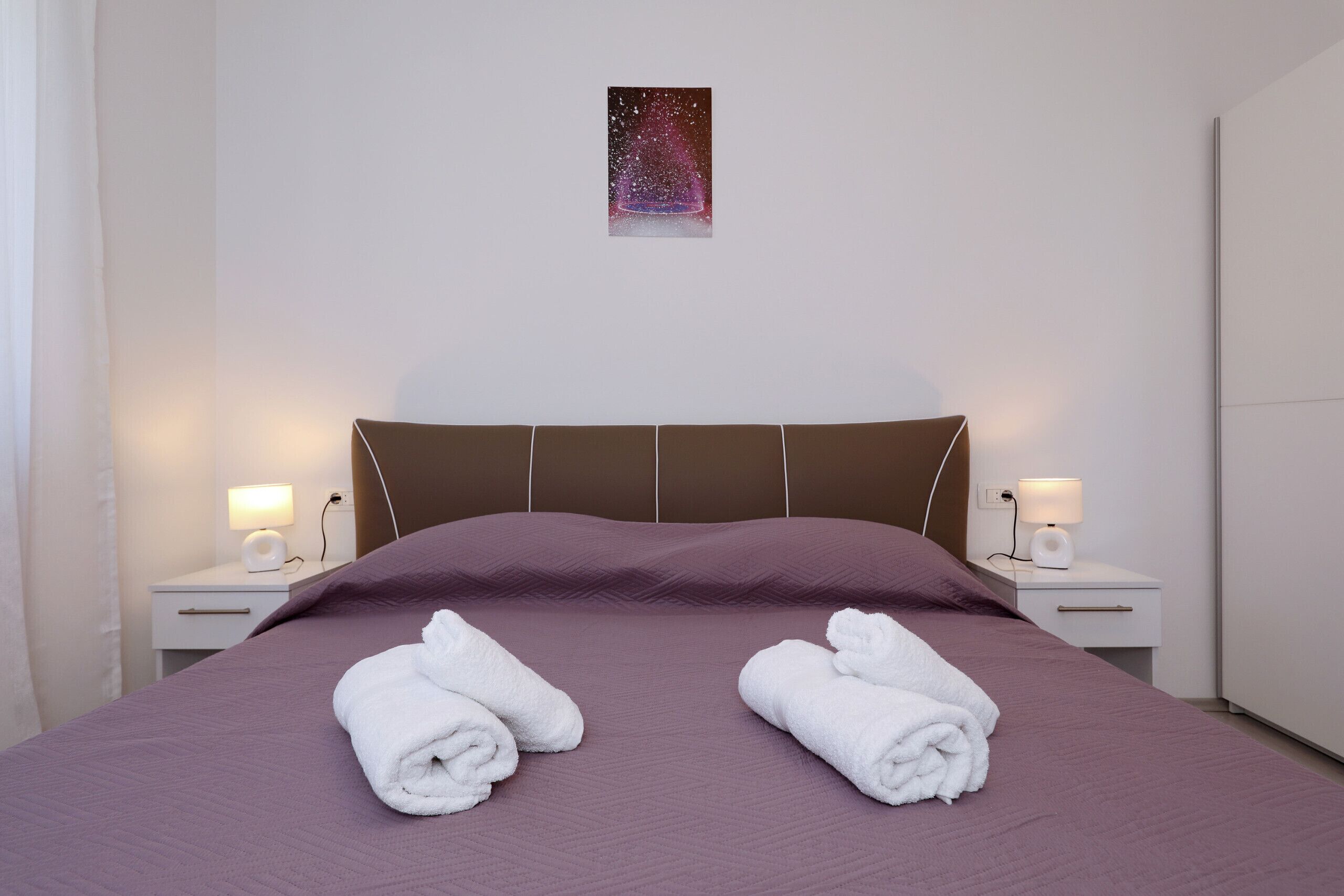Een net opgemaakt bed met twee opgerolde handdoeken, een nachtkastje met een lamp, en een aan de muur hangend schilderij.