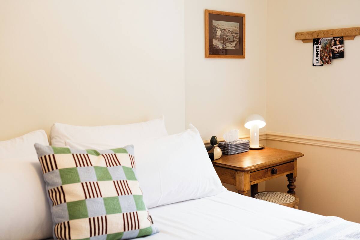 A neatly made bed with a checkered pillow, a bedside table with a lamp, a framed picture on the wall, and a shelf with books and a sign.