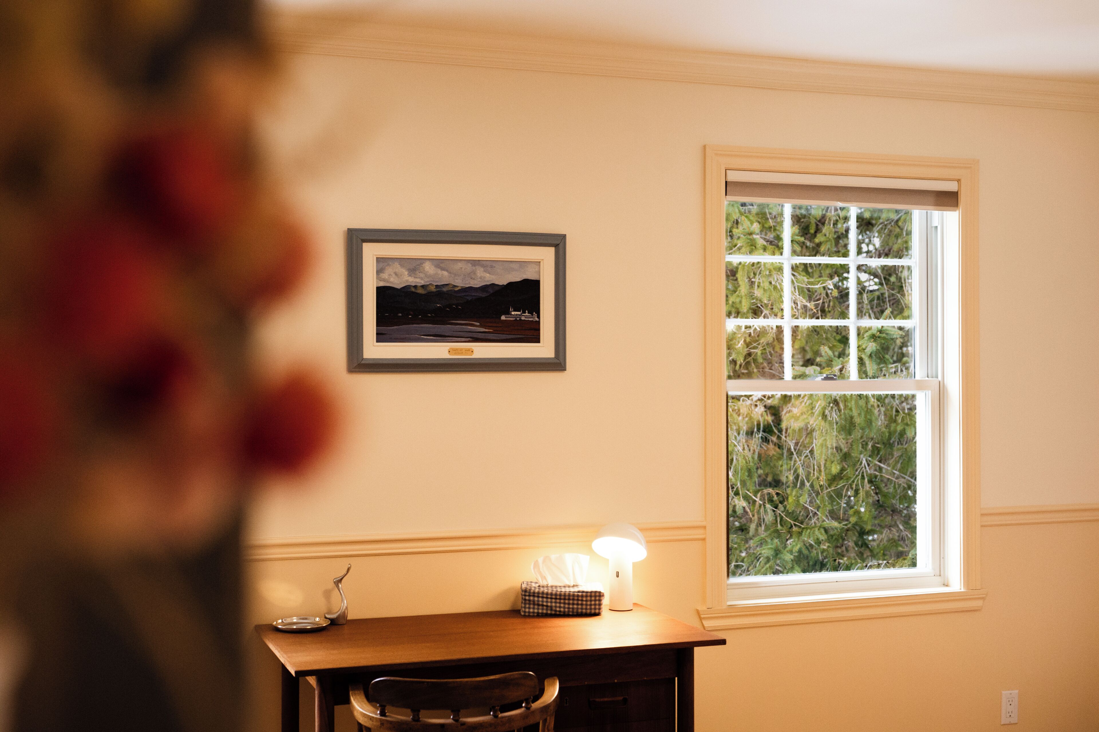 A room with a wooden desk, a framed picture on the wall, a window with a view of greenery, and a lamp on the desk.