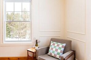 Ein Eckzimmer mit Fenster, ein Sessel mit gemustertem Kissen, ein kleiner Tisch mit Weinflasche und Glas sowie ein Korbstuhl.