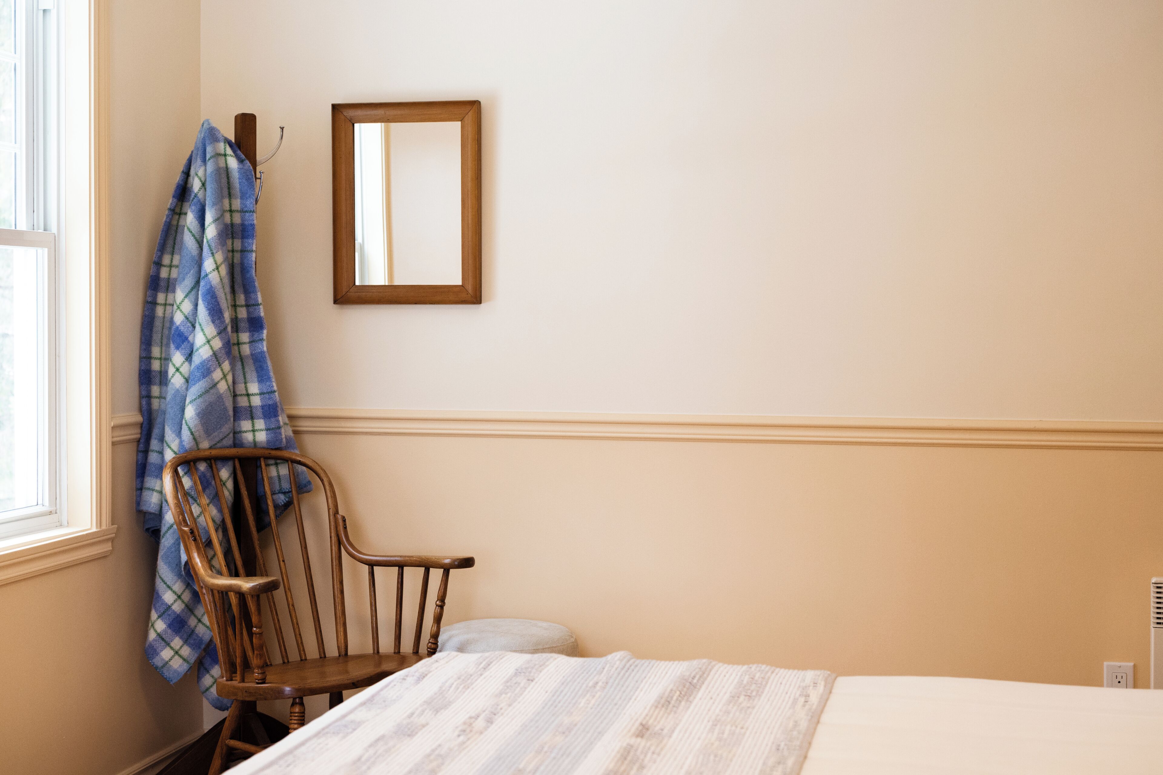 A bedroom with a bed, a wooden chair, a window, and a wall-mounted mirror.