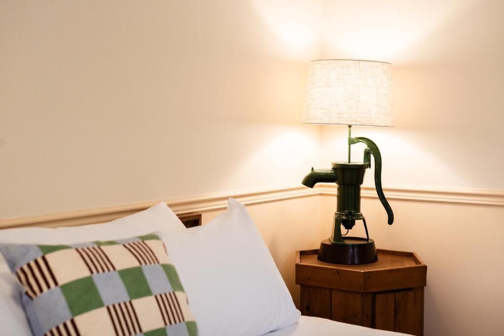 Ein Bett mit einem karierten Kopfkissen, eine grüne Lampe auf einem hölzernen Nachttisch und eine Wand mit strukturierter Oberfläche.