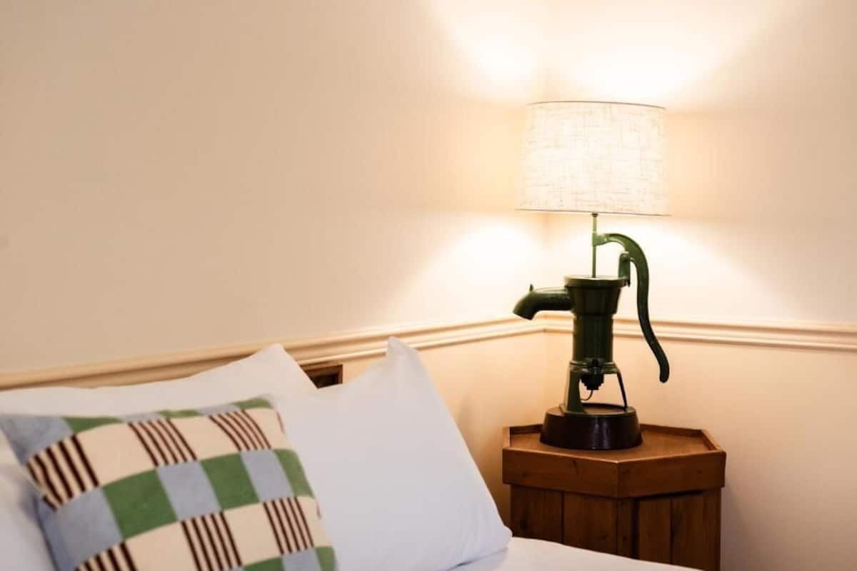 Ein Bett mit einem karierten Kopfkissen, eine grüne Lampe auf einem hölzernen Nachttisch und eine Wand mit strukturierter Oberfläche.