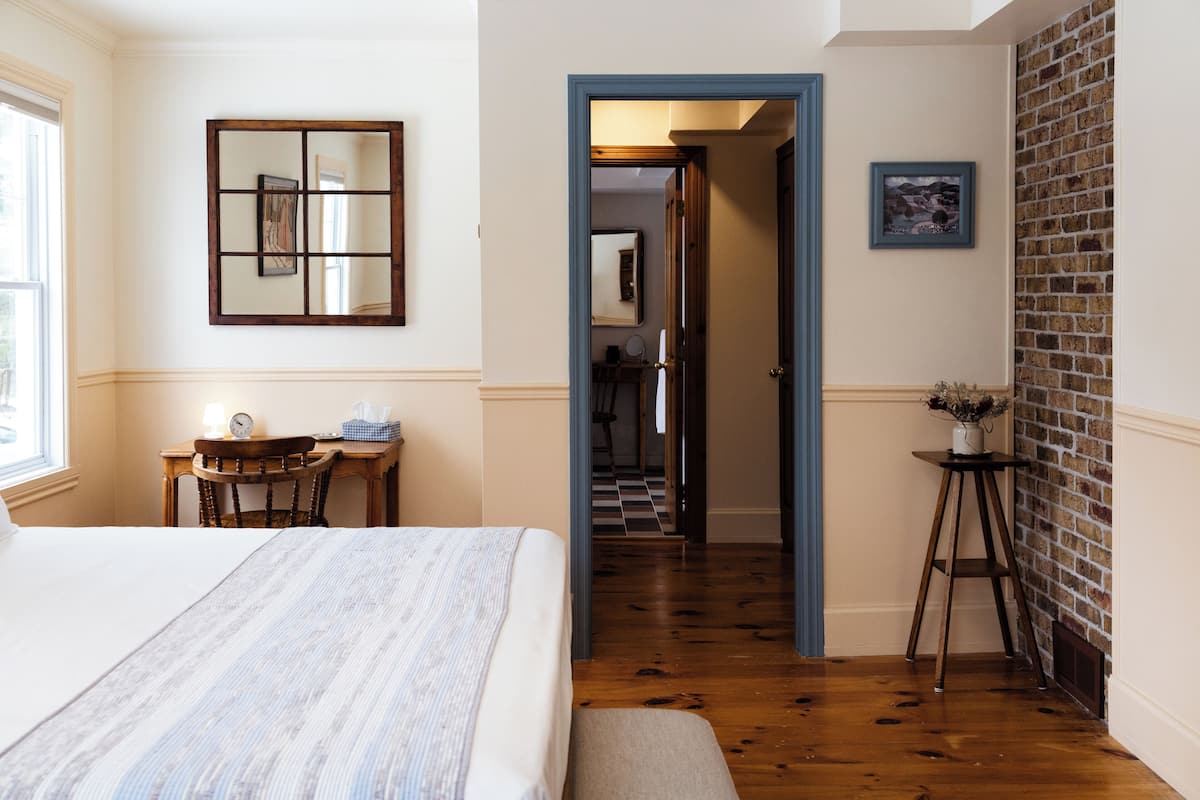A bedroom with a bed, a desk, a chair, a mirror, and a door leading to a hallway.