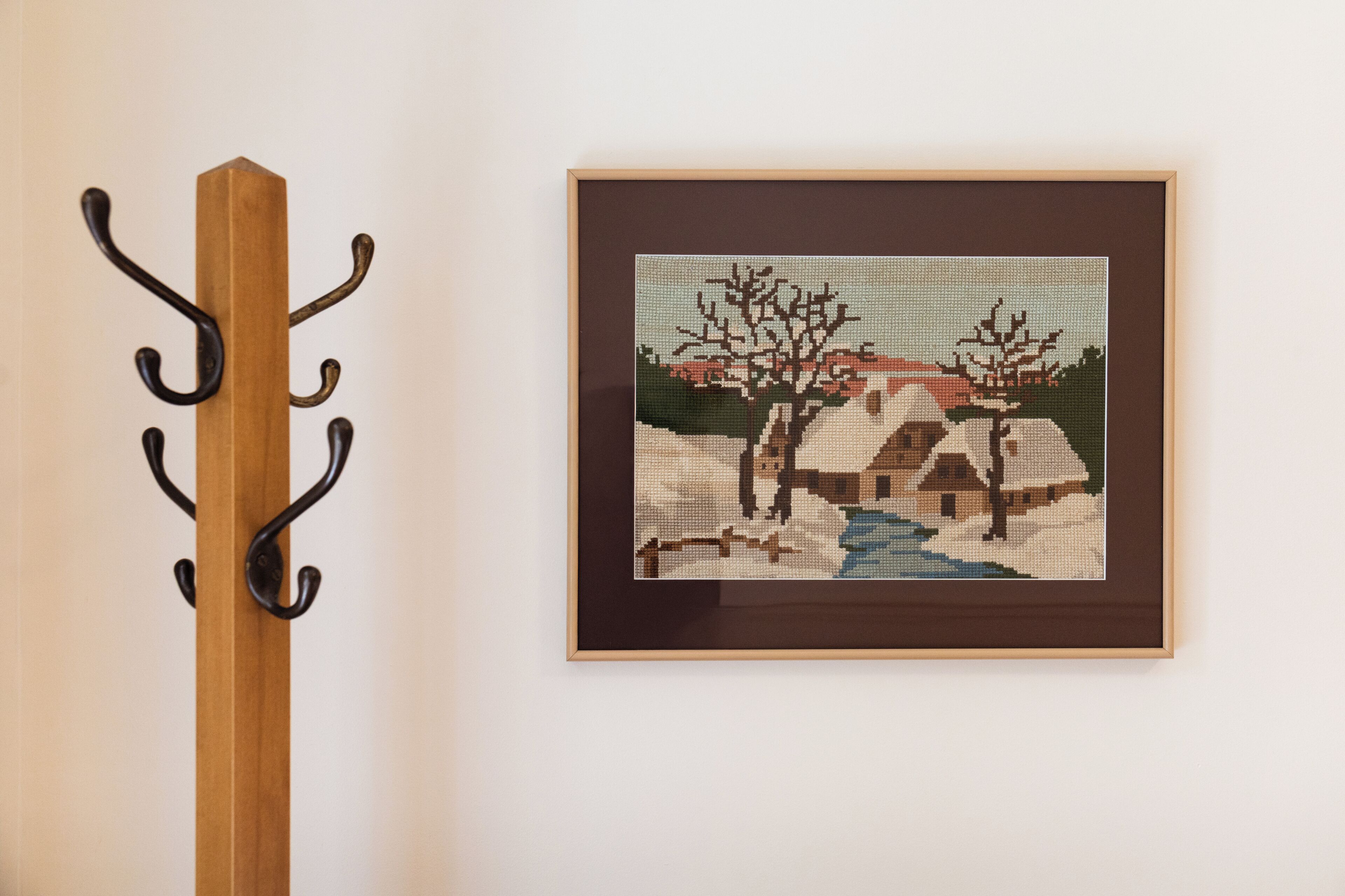 A framed cross-stitch artwork on a wall, a wooden coat rack with hooks, and a plain wall.