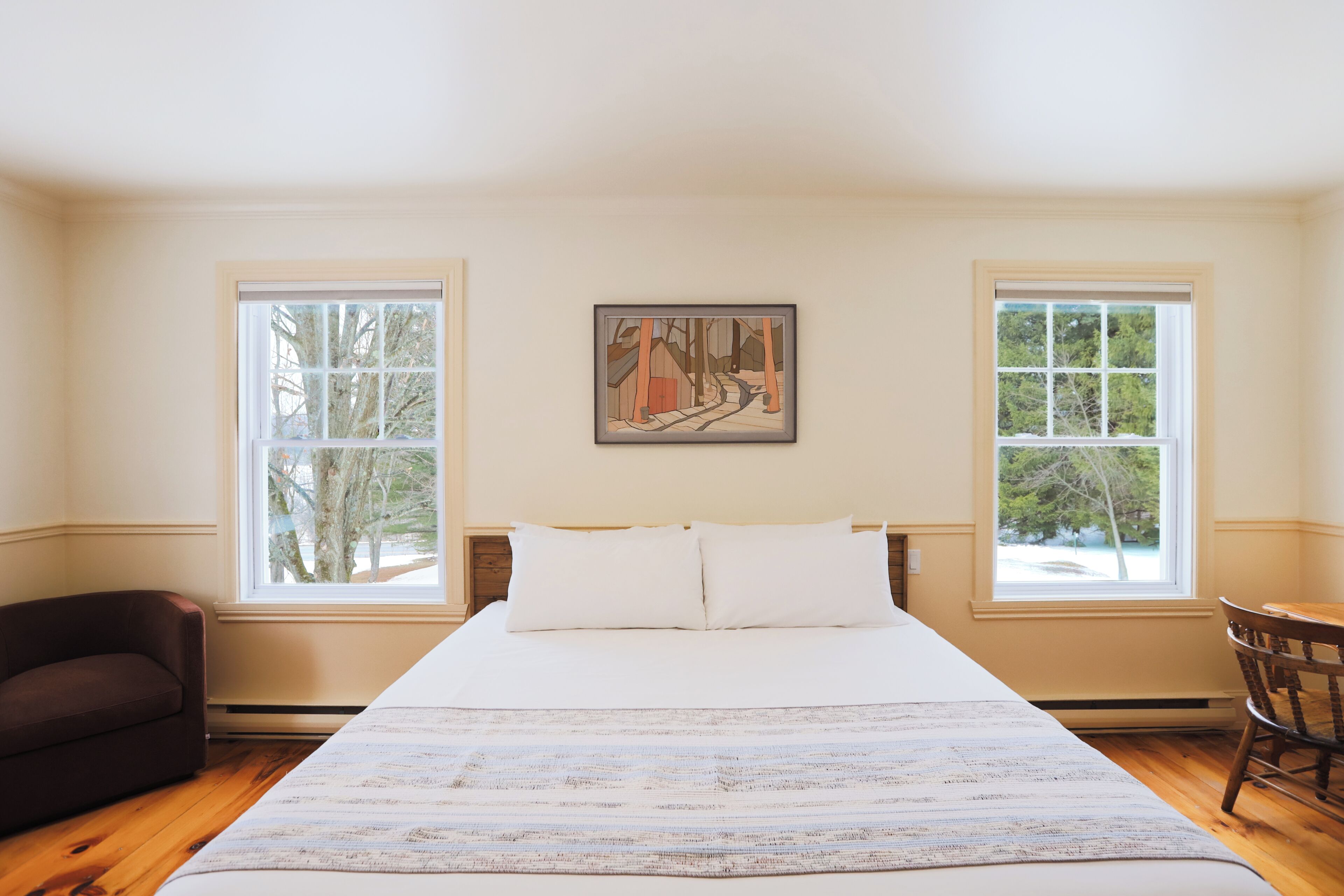 A bedroom with a large bed, two windows, a painting, a chair, and a wooden floor.