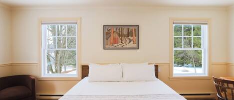 A bedroom with a large bed, two windows, a painting, a chair, and a wooden floor.