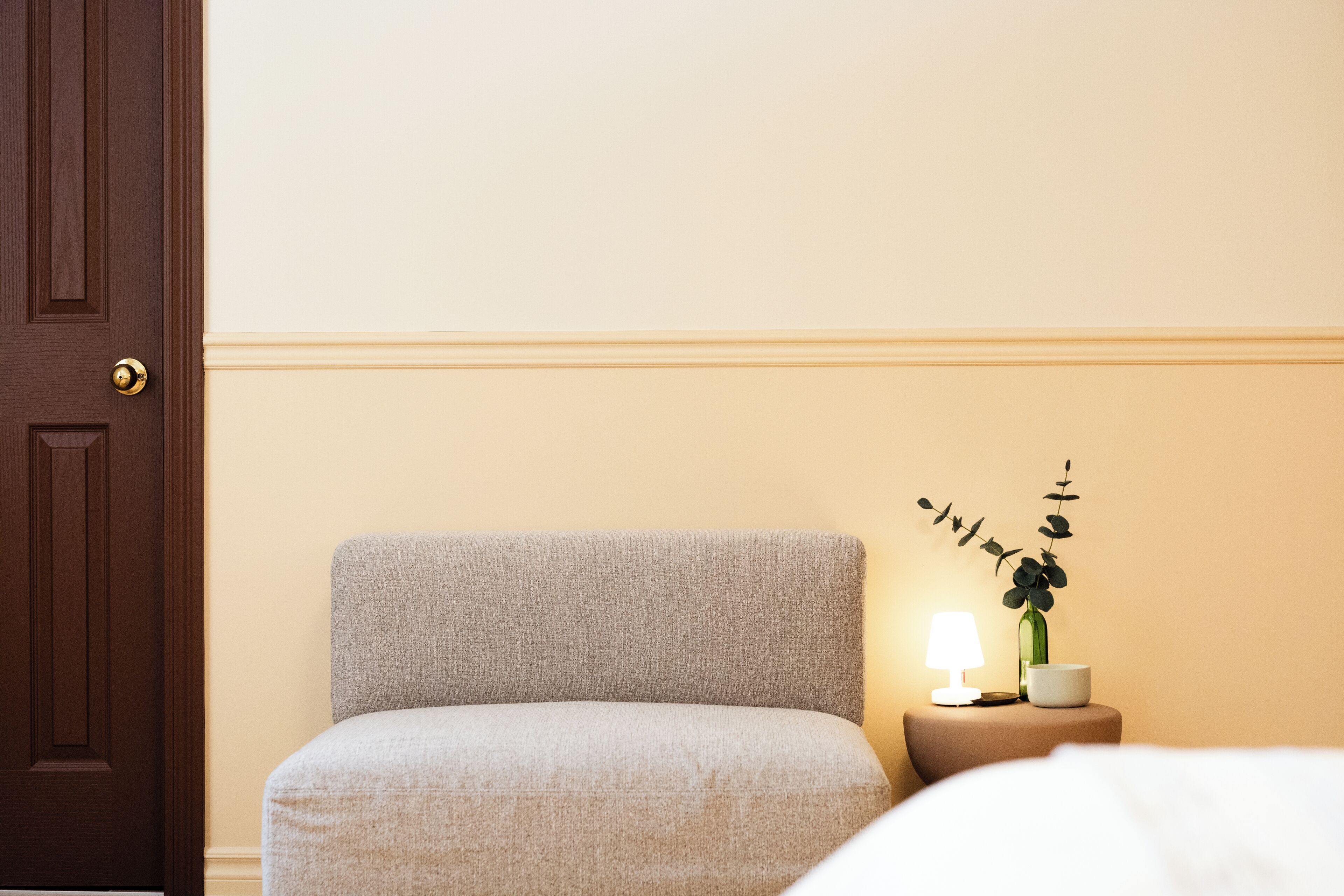 Une pièce avec une porte en bois foncé, un fauteuil rembourré de couleur claire, une table de chevet avec une lampe, un vase avec une plante et un bol blanc.