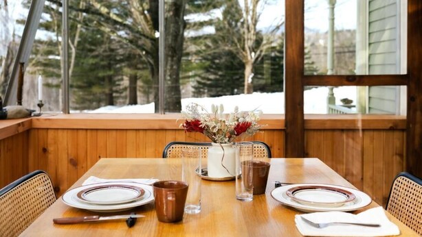 A wooden dining table with plates, cutlery, and a vase of flowers, set against a window with a view of trees. - Maison Hôte (Orford)