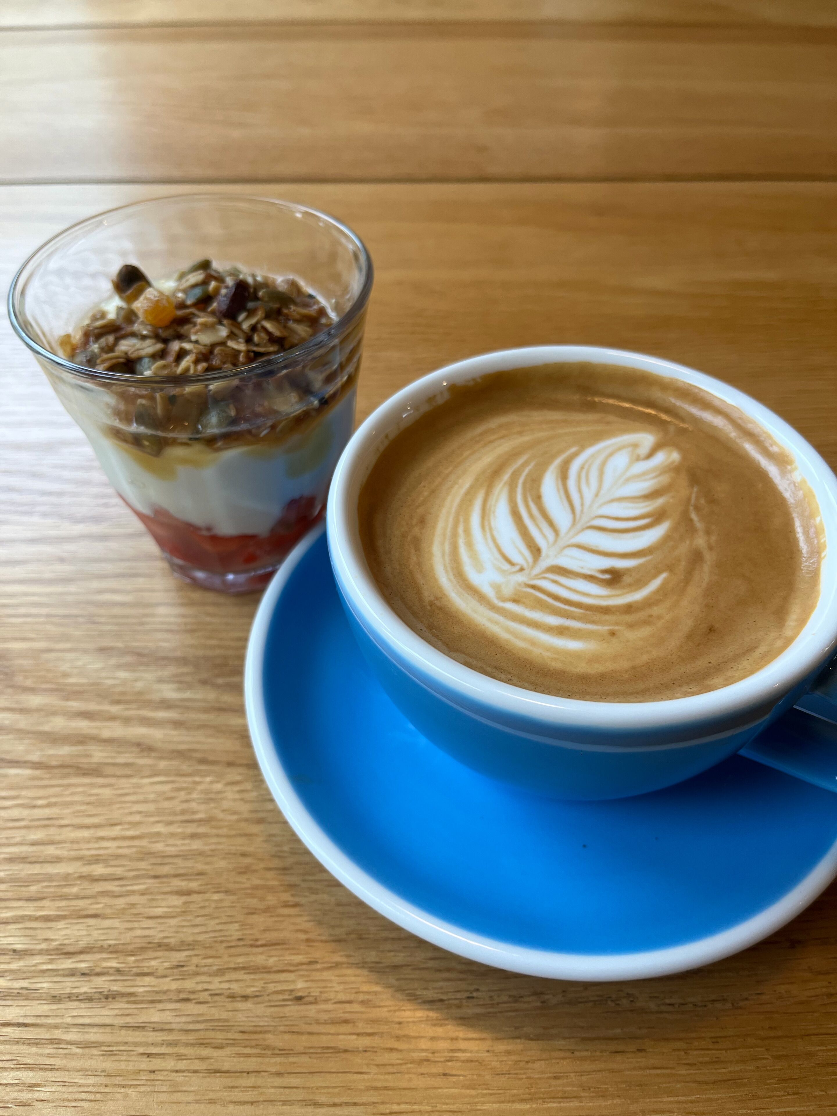 Una taza de café con decoración de leche, un vaso de parfait de yogur y una mesa de madera.