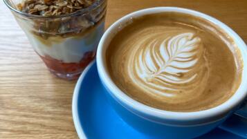 Una taza de café con decoración de leche, un vaso de parfait de yogur y una mesa de madera.