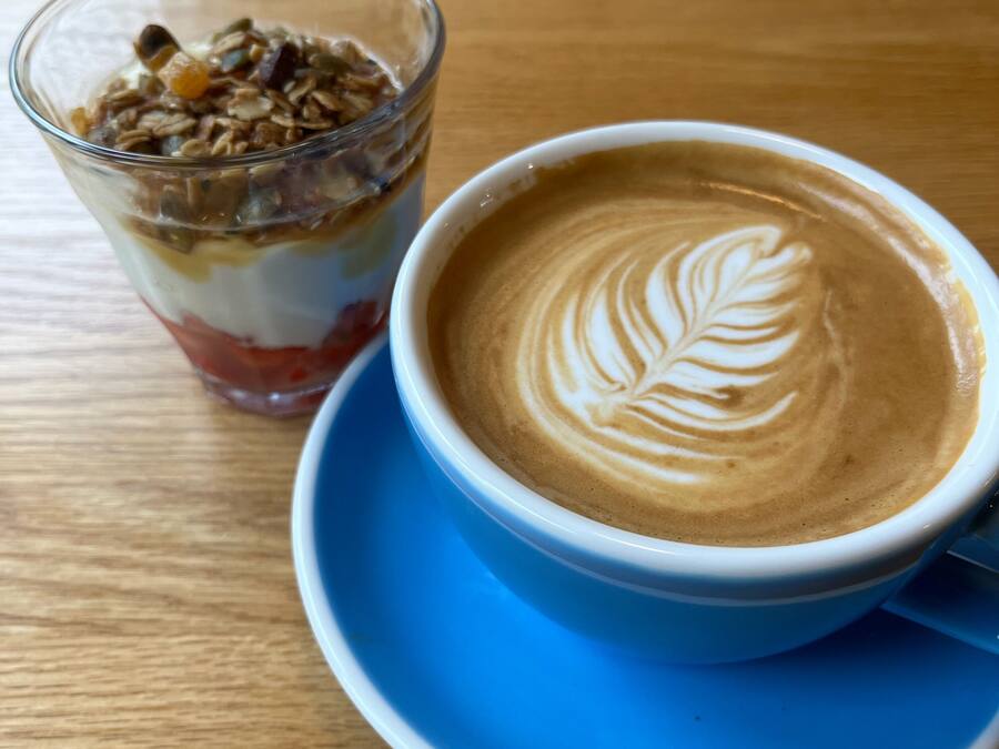 Una taza de café con decoración de leche, un vaso de parfait de yogur y una mesa de madera.