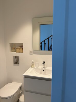 A bathroom with a white sink, a mirror, a toilet, and a small shelf with toiletries. - Maison Soleil sea view Cassis (Cassis)
