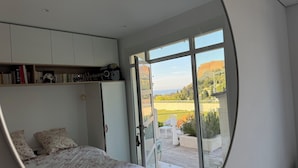 Ein Schlafzimmer mit Bett, Schrank, Regal mit Büchern und Gitarre, sowie eine große Schiebeglasdoor, die zu einem Außenbereich mit Blick auf einen Hügel und Bäume führt.