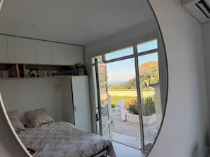 A bedroom with a bed, a wardrobe, a shelf with books and a guitar, and a large sliding glass door leading to an outdoor area with a view of a hill and trees. - Maison Soleil sea view Cassis (Cassis)
