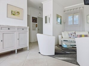 Ein Wohnzimmer mit einem weißen Sofa, einem Glastisch, einer weißen Kommode und einer Tür zu einem weiteren Raum.