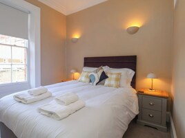 A neatly made bed with white linens and folded towels, a wooden headboard, bedside lamps, and a window with blinds.