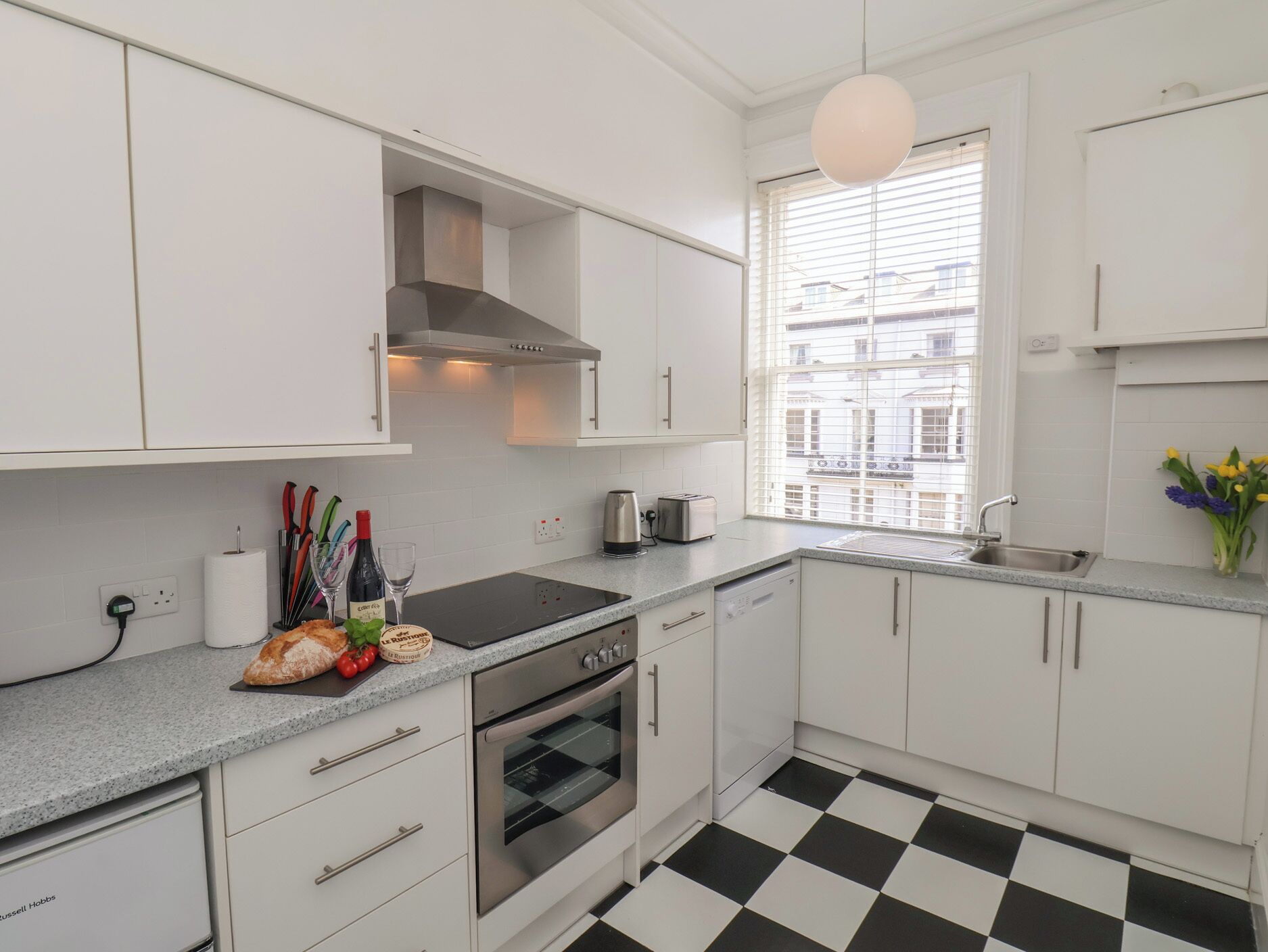 A modern kitchen with white cabinets, a black and white checkered floor, a stainless steel oven, and a window with blinds.