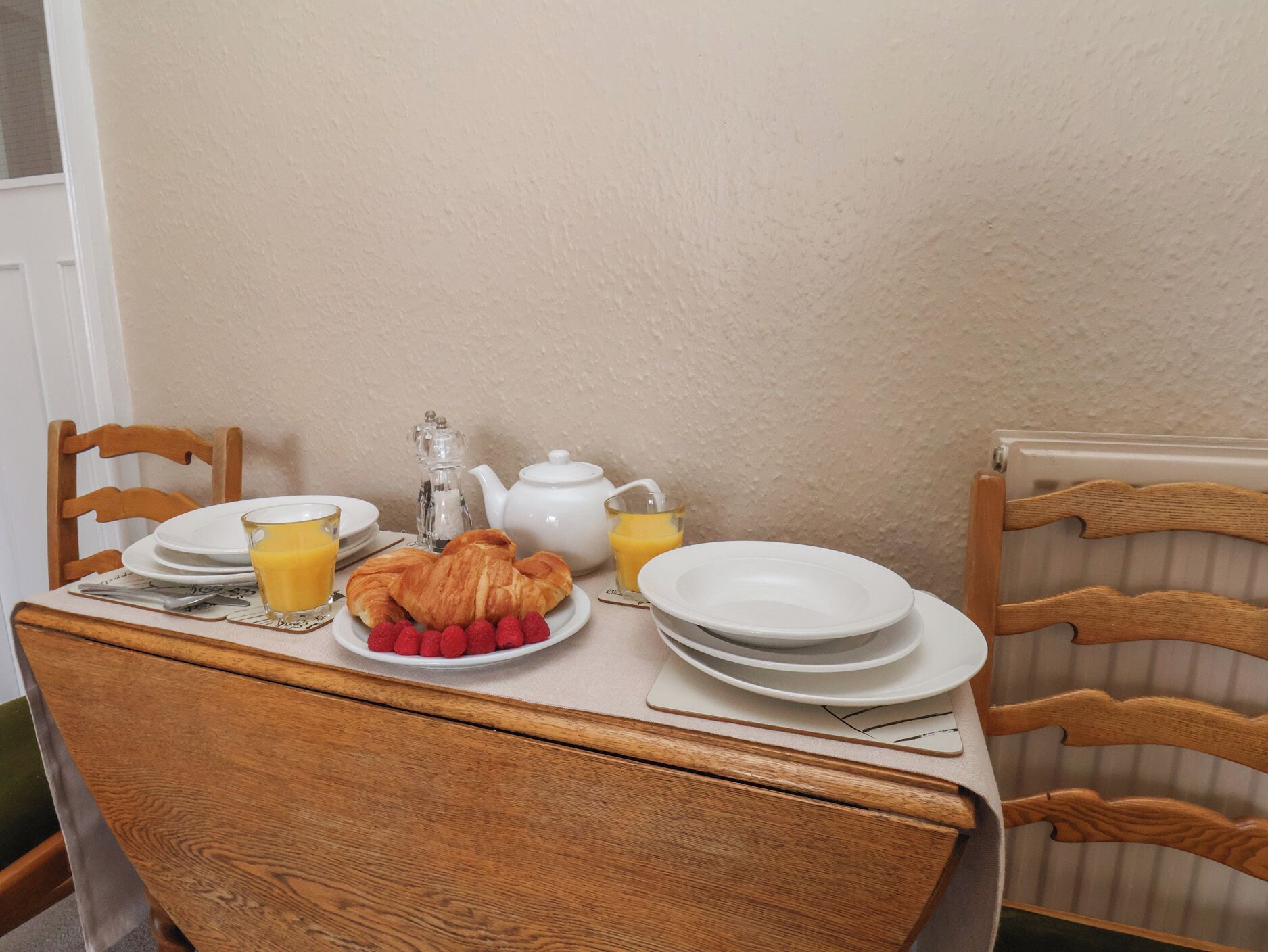 Una mesa de desayuno con cruasanes, frambuesas, una tetera y un vaso de jugo de naranja.