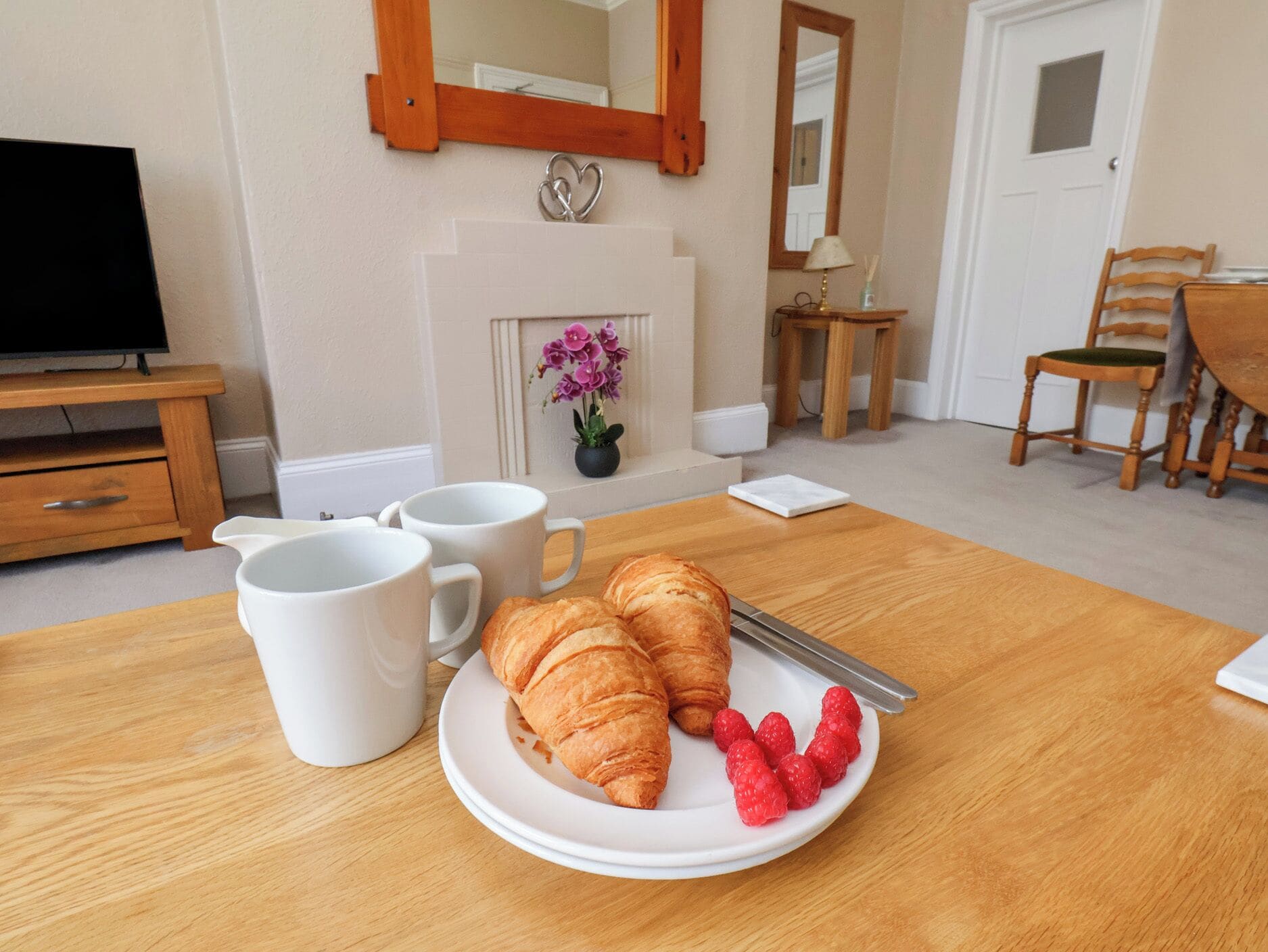 A dining table with croissants and raspberries, two white mugs, a fireplace with flowers, a television, and a mirror.